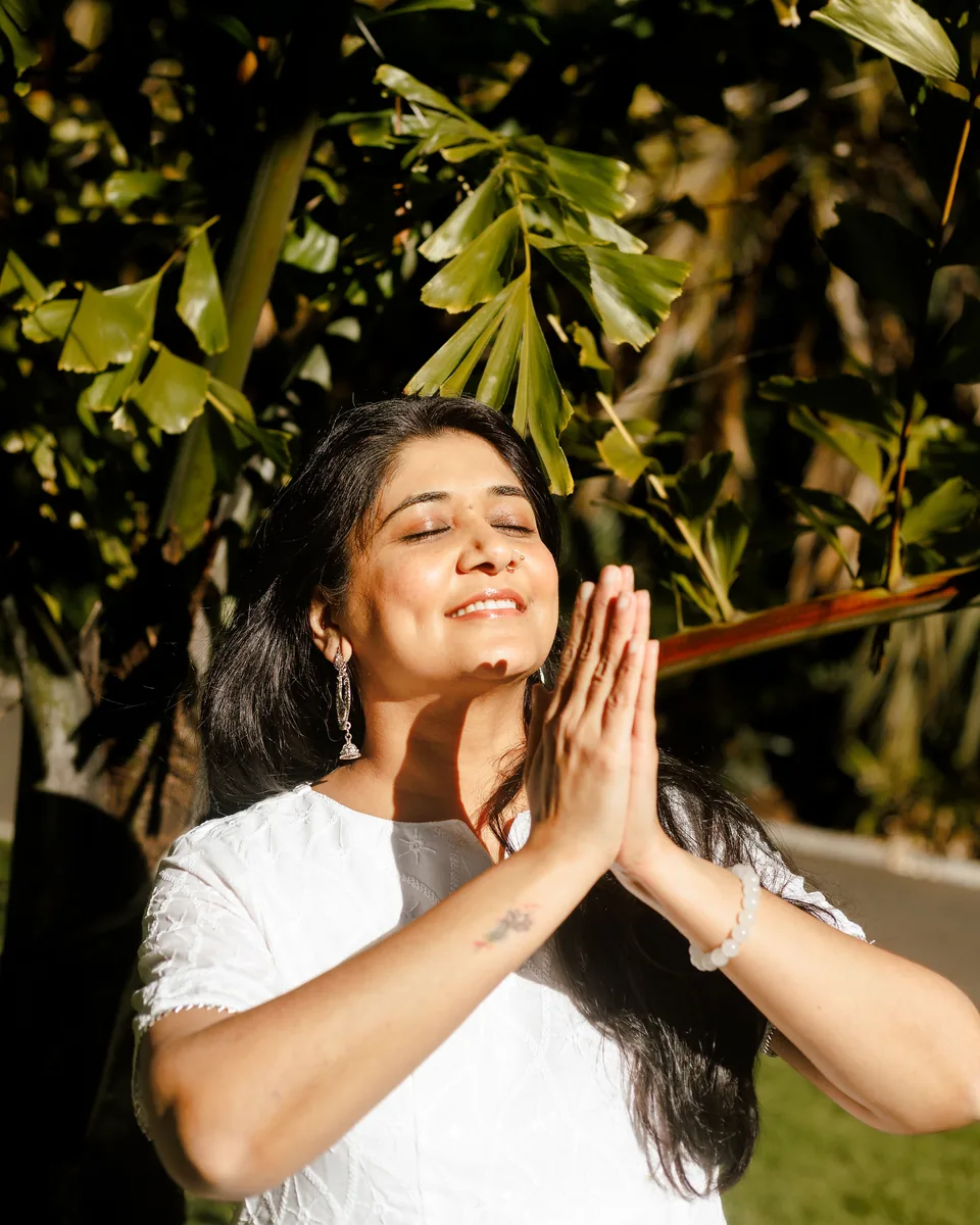 Harsiddhi Pancholi with her hands pressed together in a calm moment, sunlight through green leaves behind her