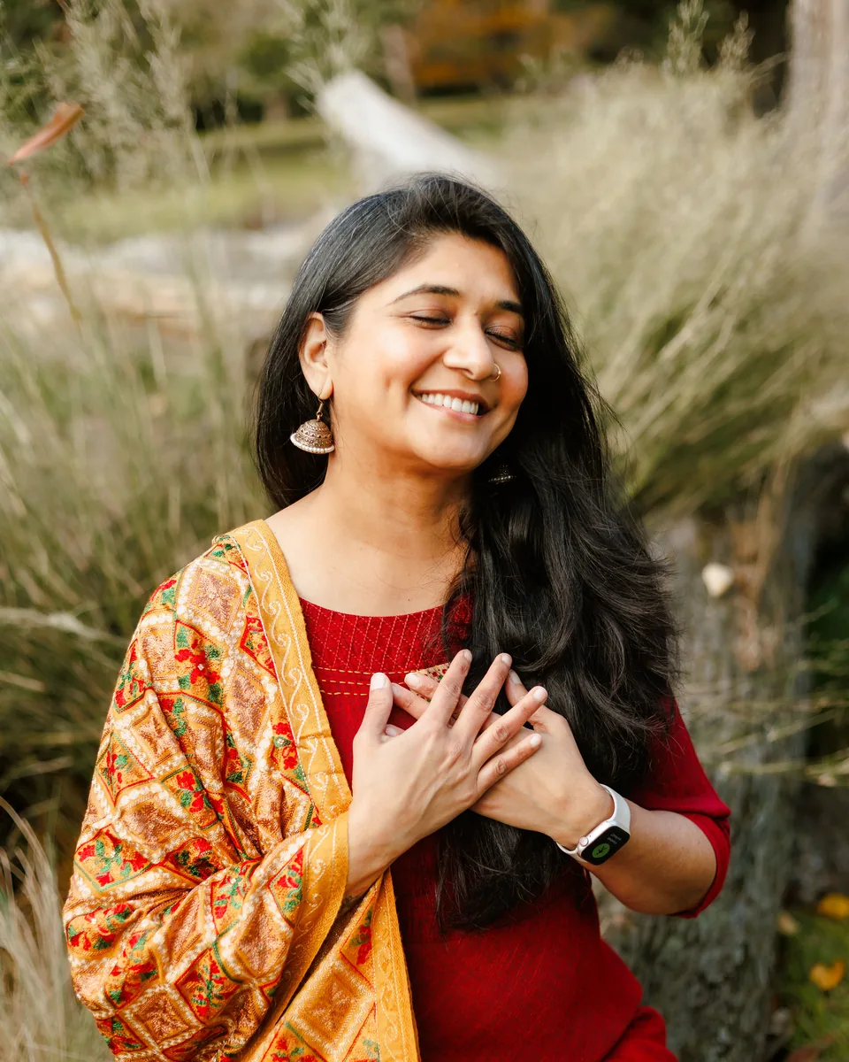 Harsiddhi Pancholi smiling with her hands on her heart, standing among tall grasses in warm light